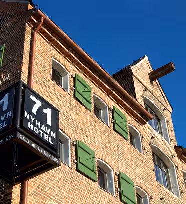 Exteriör på 71 Nyhavn Hotel, en historisk tegelbyggnad med gröna fönsterluckor och en klarblå himmel i bakgrunden. En svart skylt som visar hotellets namn och nummer är väl synlig i hörnet.
