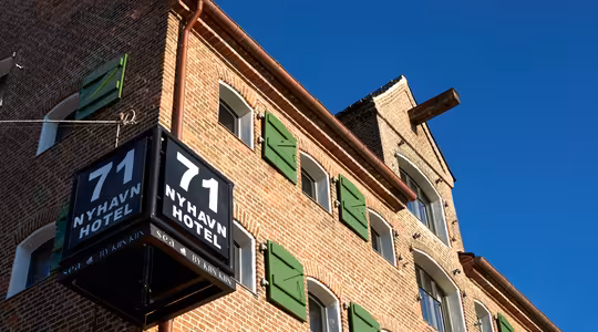 Exteriör på 71 Nyhavn Hotel, en historisk tegelbyggnad med gröna fönsterluckor och en klarblå himmel i bakgrunden. En svart skylt som visar hotellets namn och nummer är väl synlig i hörnet.