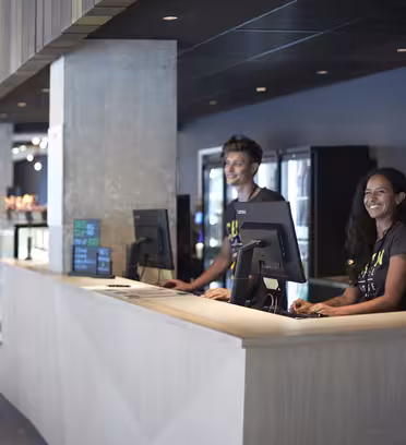 Två personer som ler bakom en modern reception utrustad med datorer. Rummet är elegant med mörka väggar och minimalistiska designelement. I bakgrunden finns montrar och hyllor.