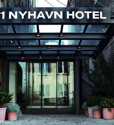 Entré till 71 Nyhavn Hotel med glastak. Fasaden har tegelväggar på båda sidor och krukväxter kantar entrén, vilket skapar en välkomnande atmosfär.