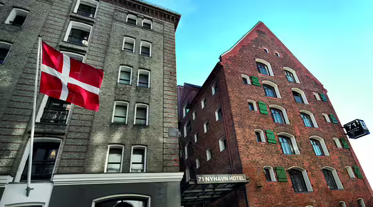 Exteriörvy av 71 Nyhavn Hotel, med en tegelbyggnad med gröna fönsterluckor och en dansk flagga vajande framför. En klarblå himmel fungerar som bakgrund och framhäver de arkitektoniska detaljerna i den historiska strukturen.