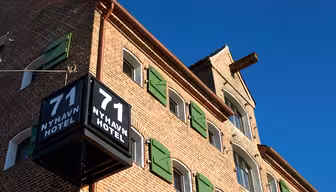 Tegelbyggnad med gröna fönsterluckor och en svart skylt som visar "71 Nyhavn Hotel" i vit text. Himlen är klar och blå, vilket förstärker byggnadens arkitektur och färger.