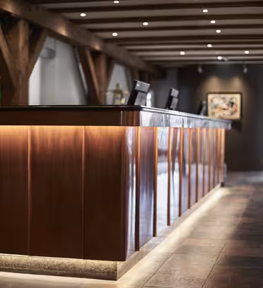 En väl upplyst hotellreception med ett långt träskrivbord och två datorstationer. Golvet är kaklat och taket har synliga träbjälkar. Målningar visas på väggarna i bakgrunden.