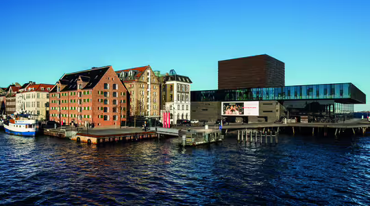 Panoramautsikt över en strand med färgglada historiska byggnader, båtar anlagda längs piren och en modern struktur i glas och tegel som sträcker sig över vattnet under en klarblå himmel.