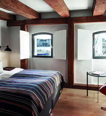Mysigt hotellrum med en dubbelsäng med ett randigt överkast, träbjälkar och välvda fönster. Ett litet runt bord med en lampa och en stol finns nära fönstret och erbjuder utsikt över en gatubild vid vattnet.