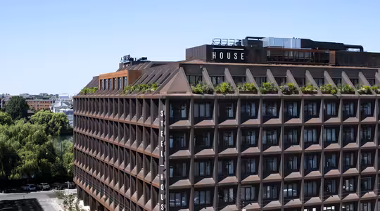 En modern flervåningsbyggnad med brun fasad och gröna växter på taket. Strukturen har ett rutmönster med stora fönster och är märkt med "STEEL HOUSE" skyltning. Träd och andra byggnader syns i bakgrunden.