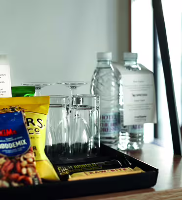 En bricka på ett bord rymmer en flaska rött vin, fyra tomma glas, två vatten på flaska och diverse snacks inklusive nötter och choklad. Bakom brickan finns en lapp till gästerna, och bakgrunden har ett fönster med mjukt, naturligt ljus.