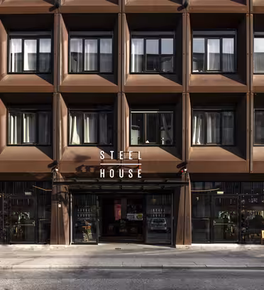 Gatuvy av en byggnadsfasad med modern, geometrisk design. Utsidan är gjord av bruna paneler och har en skylt med texten "STEEL HOUSE". Stora fönster syns och ett par bänkar är placerade nära entrén.