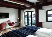 Ett mysigt hotellrum med träbjälkar har en säng med en randig filt, två bruna stolar, en tv och ett skrivbord. Stora välvda fönster erbjuder utsikt över en strand och byggnader över vattnet.
