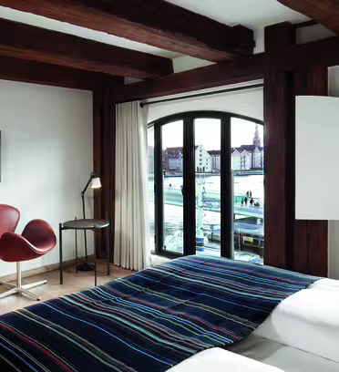 Ett mysigt hotellrum med träbjälkar har en säng med en randig filt, två bruna stolar, en tv och ett skrivbord. Stora välvda fönster erbjuder utsikt över en strand och byggnader över vattnet.