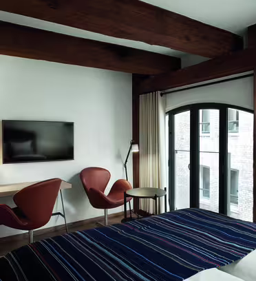 Hotellrum med synliga träbjälkar, ett randigt överkast och ett fönster som visar en tegelstensexteriör. Det finns en väggmonterad TV, ett skrivbord med två orangea stolar, ett litet runt bord och en golvlampa bredvid fönstret.