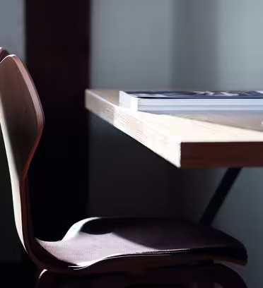 En minimalistisk arbetsyta med en trästol bredvid ett träskrivbord. Solljus lyser mjukt upp ett magasin på skrivbordet. Inställningen förmedlar en ren och fridfull atmosfär.