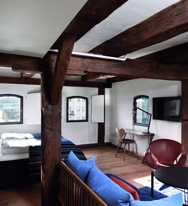 Mysigt rum i loftstil med stora träbjälkar, en säng, soffa, litet runt bord och läderstol. Ett skrivbord med en stol och en monterad TV finns bredvid välvda fönster med utsikt över staden. Trägolv förstärker den varma atmosfären.