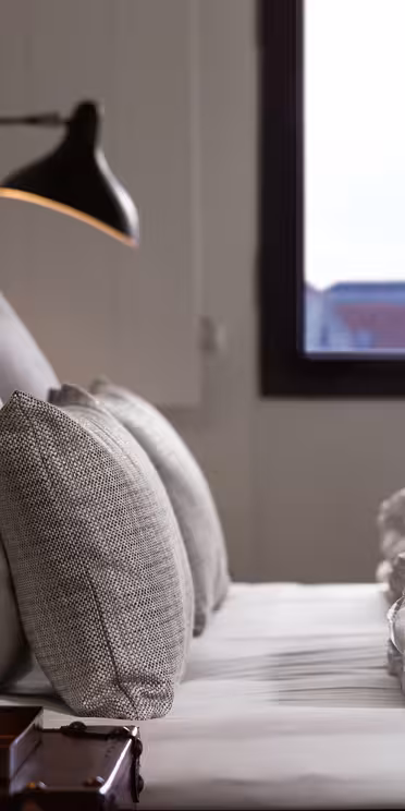 Närbild av en snyggt bäddad säng med fyra kuddar och fräscha vita sängkläder. Ett nattduksbord i trä med bricka och två väggmonterade lampor syns. Ett fönster erbjuder utsikt över avlägsna byggnader och en spira mot en klar himmel.