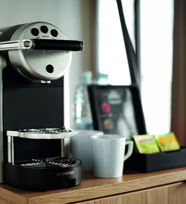 En Nespresso-maskin står på en bänkskiva i trä, med två vita kaffemuggar bredvid. I bakgrunden finns en bricka med en vattenflaska och olika tepåsar.