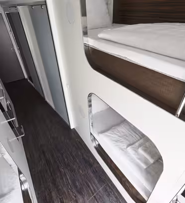 Interiörvy av en modern, smal kapselhotellhall med vita väggar och mörka trägolv. Våningssängar är integrerade i väggarna med vita sängkläder. Stängda dörrar syns i slutet av korridoren.