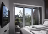 Modernt hotellrum med snyggt bäddad dubbelsäng. En platt-TV är monterad på den vita väggen. Stora fönster från golv till tak erbjuder utsikt över grönska och klar himmel utanför, vilket låter naturligt ljus fylla utrymmet.