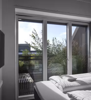 Modernt hotellrum med snyggt bäddad dubbelsäng. En platt-TV är monterad på den vita väggen. Stora fönster från golv till tak erbjuder utsikt över grönska och klar himmel utanför, vilket låter naturligt ljus fylla utrymmet.