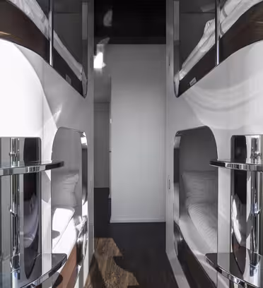 En modern kapselhotellinteriör med eleganta vita och metalliska ytor. Våningssängar är inriktade på båda sidor, med individuella sovkapslar med madrasser. Hallen är smal med trägolv och leder till ett rent, minimalistiskt utrymme.