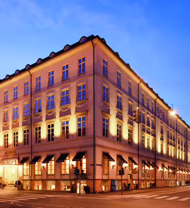 Tre våningar historisk byggnad med upplysta fönster och balkonger i skymningen. Den har rader av markiser ovanför bottenvåningen, ett sluttande tak med takkupor och ligger i ett hörn med kullerstensgator.
