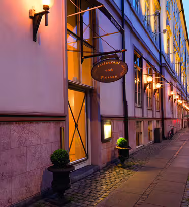 En väl upplyst gatubild med exteriören på "Restaurant von Plessen" med varma glödande ljus och en synlig skylt. Byggnaden har stora fönster och krukväxter nära dörren, med en cykel parkerad i bakgrunden.