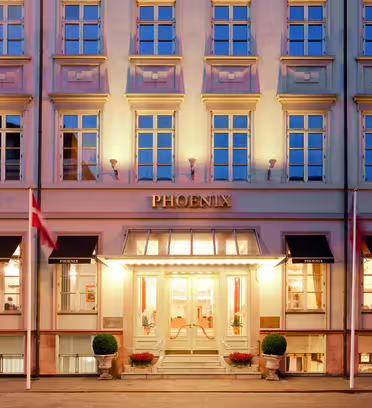 Bilden visar fasaden på Hotel Phoenix i skymningen. Byggnaden har en klassisk design med rader av upplysta fönster. Ett par flaggor flankerar entrén som är varmt upplyst och prydd med planteringskärl.