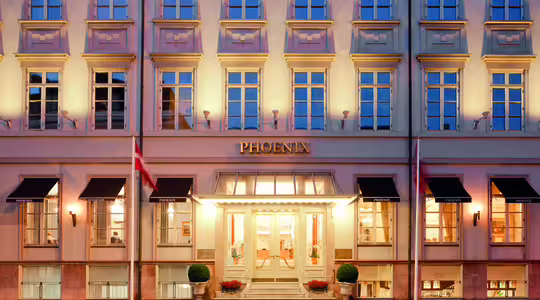 Bilden visar fasaden på Hotel Phoenix i skymningen. Byggnaden har en klassisk design med rader av upplysta fönster. Ett par flaggor flankerar entrén som är varmt upplyst och prydd med planteringskärl.