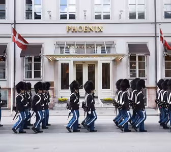En grupp uniformerade vakter i svarta hattar och blå byxor marscherar framför en byggnad med stora fönster och ordet "PHOENIX" ovanför entrén. Två röda och vita flaggor visas på vardera sidan.