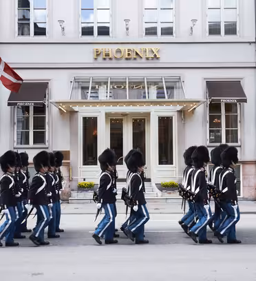En grupp uniformerade vakter i svarta hattar och blå byxor marscherar framför en byggnad med stora fönster och ordet "PHOENIX" ovanför entrén. Två röda och vita flaggor visas på vardera sidan.