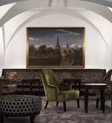 En mysig loungedel med mönstrade soffor och fåtöljer runt små bord, centrerad under en stor landskapsmålning. Rummet har vita välvda tak, eleganta vägglampetter och en mjuk matta under fötterna.