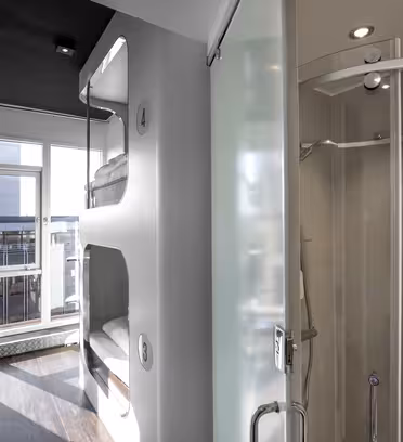 Ett modernt vandrarhemsrum med våningssängar i podstil på vänster sida och en inglasad dusch till höger. Solljus strömmar in genom stora fönster i slutet av korridoren och lyser upp den eleganta interiören.