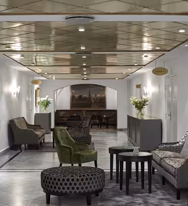 En hotellobby med eleganta sittplatser inklusive fåtöljer och en mönstrad ottoman. Taket har infälld belysning och stora tavlor pryder väggarna. Buketter av vita blommor står på sidoborden, vilket förstärker den sofistikerade atmosfären.
