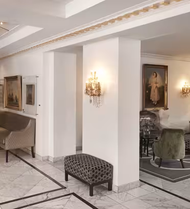 En sofistikerad hotellobby med marmorgolv och eleganta möbler. Till vänster står en lång bänk mot väggen. Den högra sidan har en sittgrupp med färgglada stolar, en matta och målningar på väggarna. Varm belysning förhöjer stämningen.