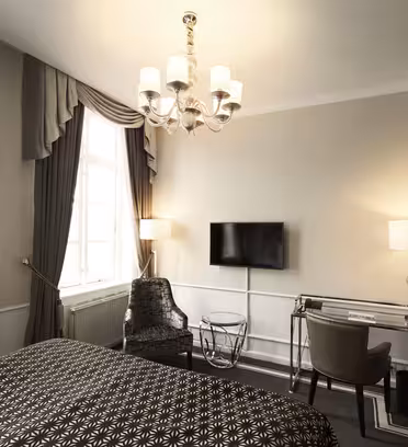 Ett elegant hotellrum har en mysig säng med mönstrat täcke, en modern fåtölj och ett elegant skrivbord i kombination med en läderstol. En platt-TV är monterad på väggen och en ljuskrona hänger i taket och lyser upp utrymmet.