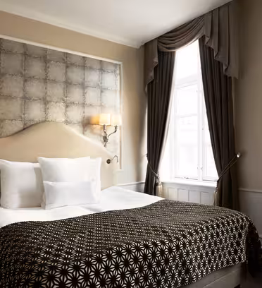Ett väl upplyst hotellrum med en stor säng med vita kuddar och en svart mönstrad filt. Väggarna är ljusa med en dekorativ panel bakom sängen. Två eleganta vägglampor lyser upp området. Ett fönster med tunga gardiner finns till höger.
