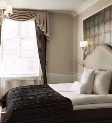 Ett mysigt hotellrum med en snyggt bäddad säng med vita kuddar och ett mörkt överkast. Väggarna är neutrala, med en stor texturerad sänggavel. Ett fönster med eleganta gardiner släpper in naturligt ljus. Ett litet skrivbord och en stol sitter i hörnet.