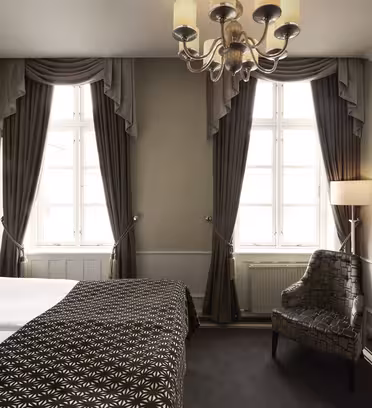 Elegant hotellrum med stora fönster, mörka gardiner, en ljuskrona och en plyschmönstrad fåtölj. En dubbelsäng med mönstrat täcke och ett litet glasbord med TV finns också, vilket skapar en mysig och sofistikerad atmosfär.