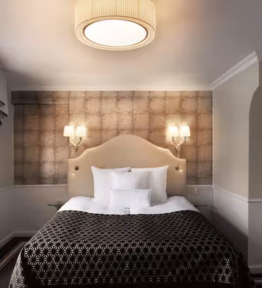 Elegant hotellrum med en lyxig dubbelsäng, mönstrat svartvitt överkast och kuddar. Sänggaveln är ljusbeige, med två lampor. Ett fönster med draperade gardiner finns till vänster och en modern taklampa ovanför. Neutrala toner genomgående.