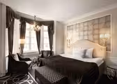 Ett lyxigt hotellrum med en stor säng med mörkrandiga sängkläder och matchande kuddar. Rummet har elegant inredning, två fåtöljer, en mönstrad ottoman, två höga fönster med gardiner, en ljuskrona och vägghängda lampor.