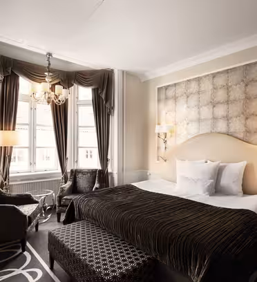 Ett lyxigt hotellrum med en stor säng med mörkrandiga sängkläder och matchande kuddar. Rummet har elegant inredning, två fåtöljer, en mönstrad ottoman, två höga fönster med gardiner, en ljuskrona och vägghängda lampor.