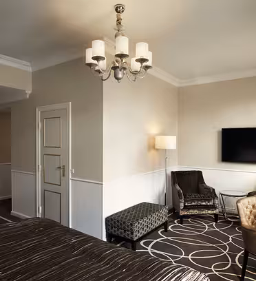 Ett hotellrum med en säng med ett mönstrat överkast i förgrunden. Det finns två fåtöljer, en bänk, golvlampa och en väggmonterad platt-TV. Rummet har beiga väggar, mörka mattor, en ljuskrona och naturligt ljus från ett fönster.