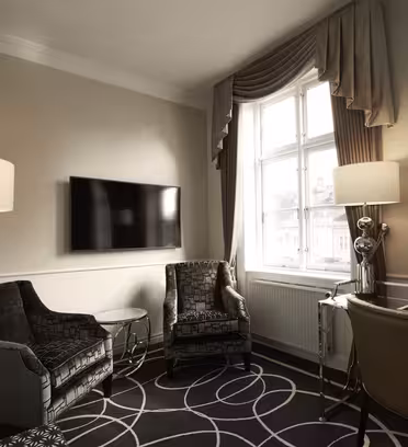 En mysig hotellrumsinredning med två mörka fåtöljer, ett sidobord i glas och en golvlampa. En platt-TV är väggmonterad. Det finns ett skrivbord och en stol till höger. Ett stort fönster med draperade gardiner tillåter naturligt ljus att lysa upp utrymmet.