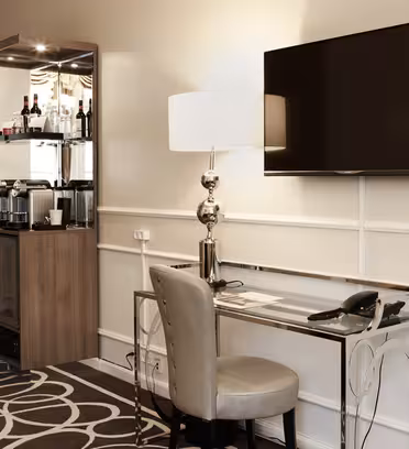 Hotellrumshörna med ett elegant skrivbord, modern stol och bordslampa. En väggmonterad platt-TV finns ovanför. En minibar med drycker sitter bredvid ett spegelskåp. Rummet har mönstrade mattor och ljusa väggar.