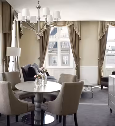Elegant hotellsvit med ett runt matbord och fyra beiga stolar, en plyschsoffa och en ljuskrona. Stora fönster med bruna draperier släpper in naturligt ljus. Mjuka neutrala toner och blommor på bordet ger värme.