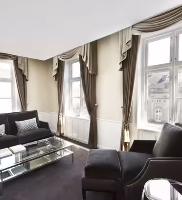 Ett stilrent vardagsrum med stora fönster och bruna draperier. Den har en svart soffa, fåtölj, soffbord i glas och två dekorativa kuddar. Lokalen är väl upplyst och har en modern, elegant design.