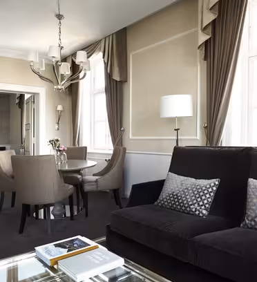 Lyxig hotellsvit med en vardagsrumsdel med en svart soffa, ett soffbord i glas och mönstrade kuddar. Det finns ett runt matbord med fyra stolar och ett angränsande sovrum synligt med en stor säng, ljuskrona och elegant inredning.