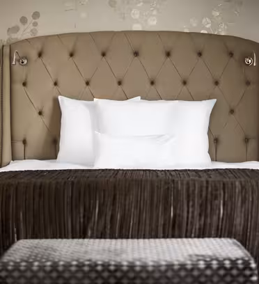 Ett hotellrum med en stor säng med en hög, tuftad sänggavel i neutral nyans. Sängen är prydd med vita kuddar och en brun plyschfilt. Två matchande sängbord och lampor flankerar sängen, och väggarna har ett subtilt blommönster.