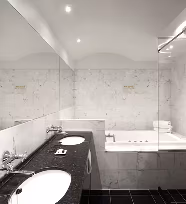 Ett modernt badrum med marmorväggar har ett handfat med dubbla handfat och ett stort badkar med jacuzzi. Det finns en bred spegel ovanför diskbänkarna och utrymmet är väl upplyst med infälld belysning.