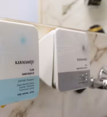 Närbild av två fyrkantiga dispensrar monterade på en badrumsvägg. Den vänstra dispensern har "Karmameju Flow Hand Wash 02" märkt i blått och vitt. Den högra dispensern är något suddig. De är monterade ovanför ett handfat i marmor med silverblandare.
