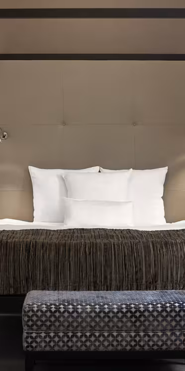Ett elegant sovrum med en modern himmelssäng med vita sängkläder och bruna dekorativa kuddar. Två väggmonterade lampor flankerar sängen på en beige accentvägg. En mönstrad bänk sitter vid fotändan av sängen på ett heltäckningsmatta golv.